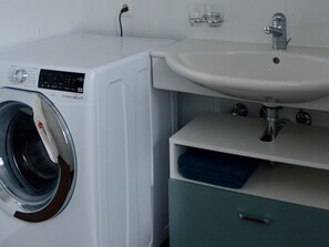 A white washing machine with a control panel, a sink with a faucet, and a blue towel on a shelf. - Chasa Rachögna, (Scuol) by Interhome (Scuol)