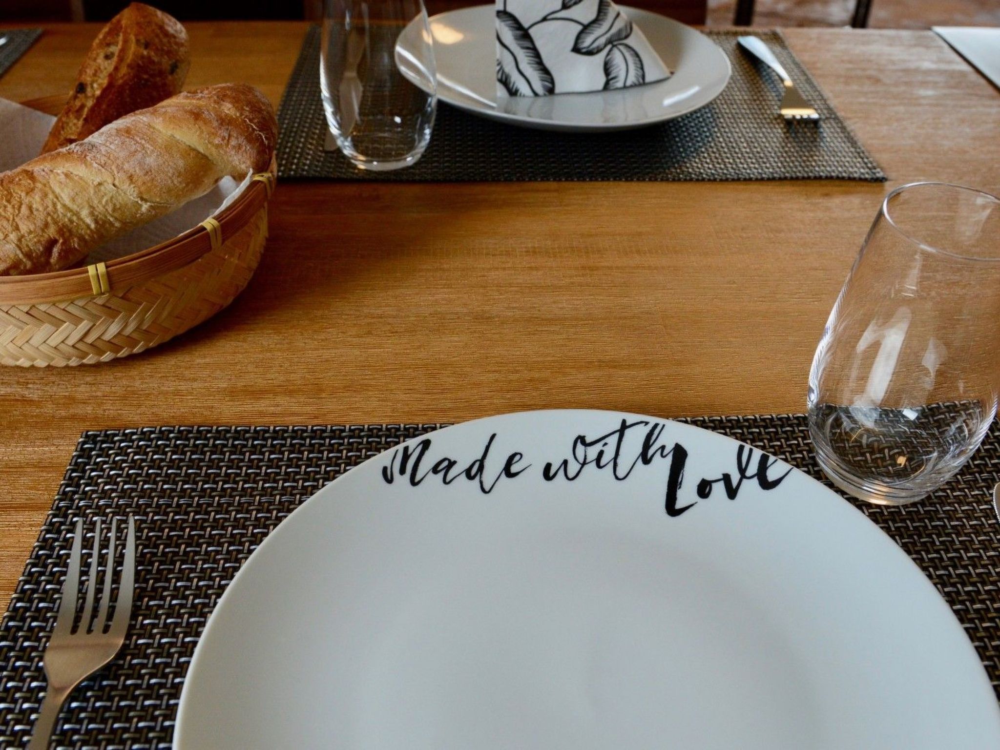 Une table à manger avec une assiette blanche, un panier de pain, un verre d’eau et une fourchette.