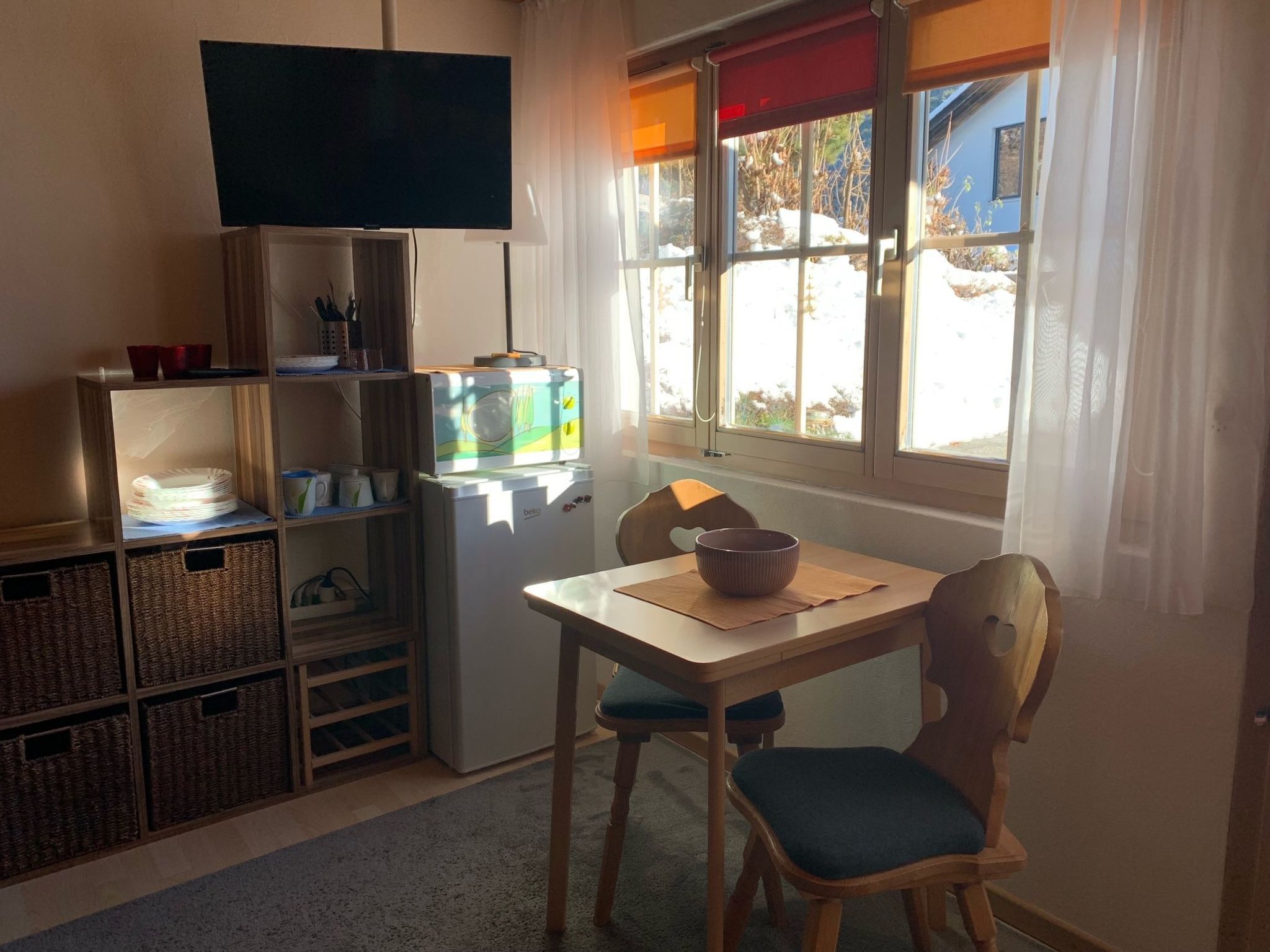 Une petite cuisine équipée d’un réfrigérateur, d’un four à micro-ondes et d’un téléviseur placé au-dessus. On y trouve également une table à manger avec des chaises et un saladier.