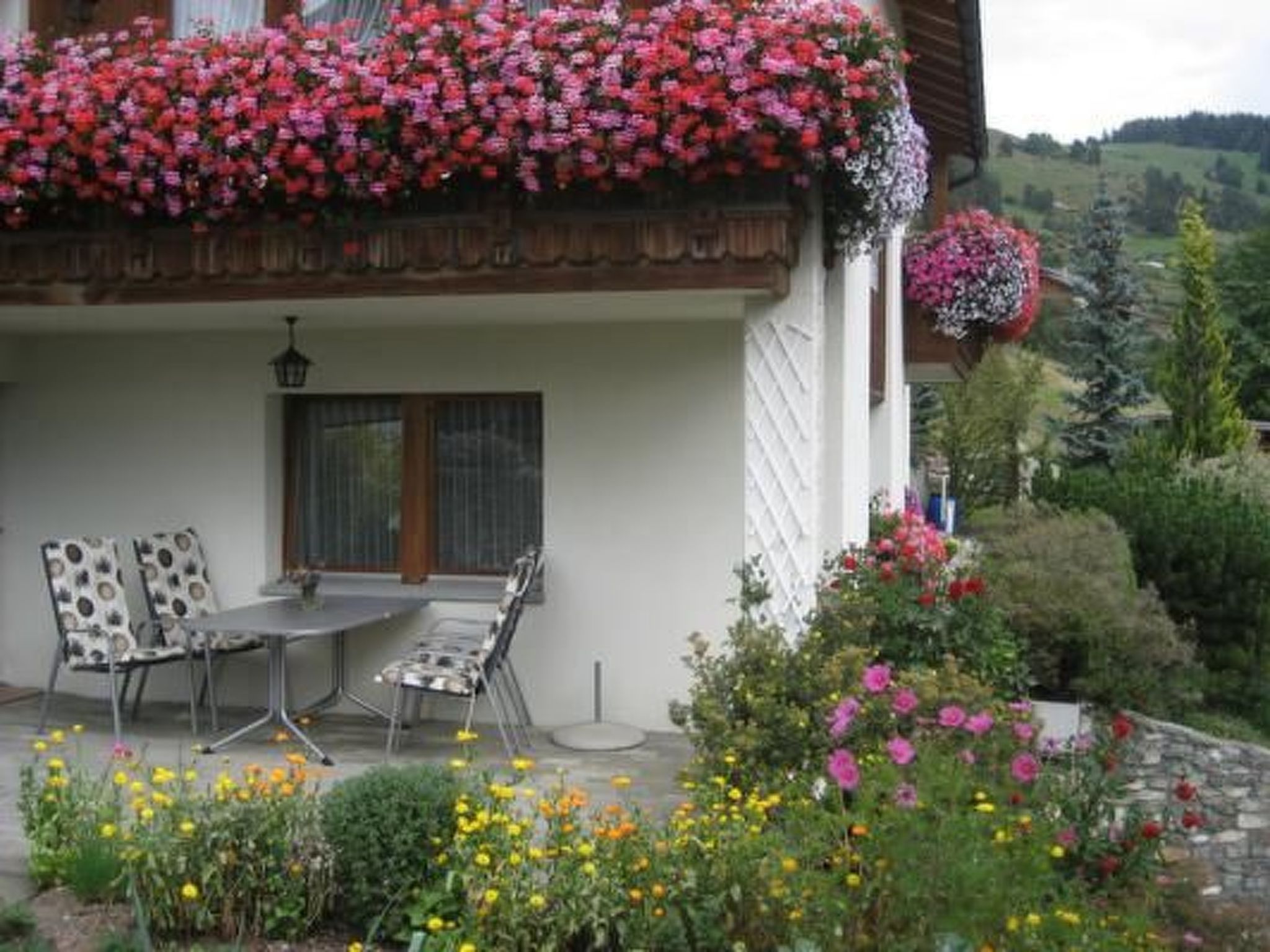 A house with a balcony, a dining table with chairs, and a garden with various flowers.
