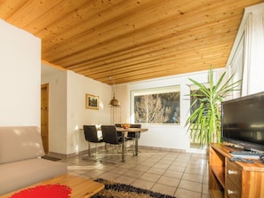 A modern living room with a wooden ceiling, a dining area with chairs and a table, a TV on a wooden stand, and a large window with a view of trees.
