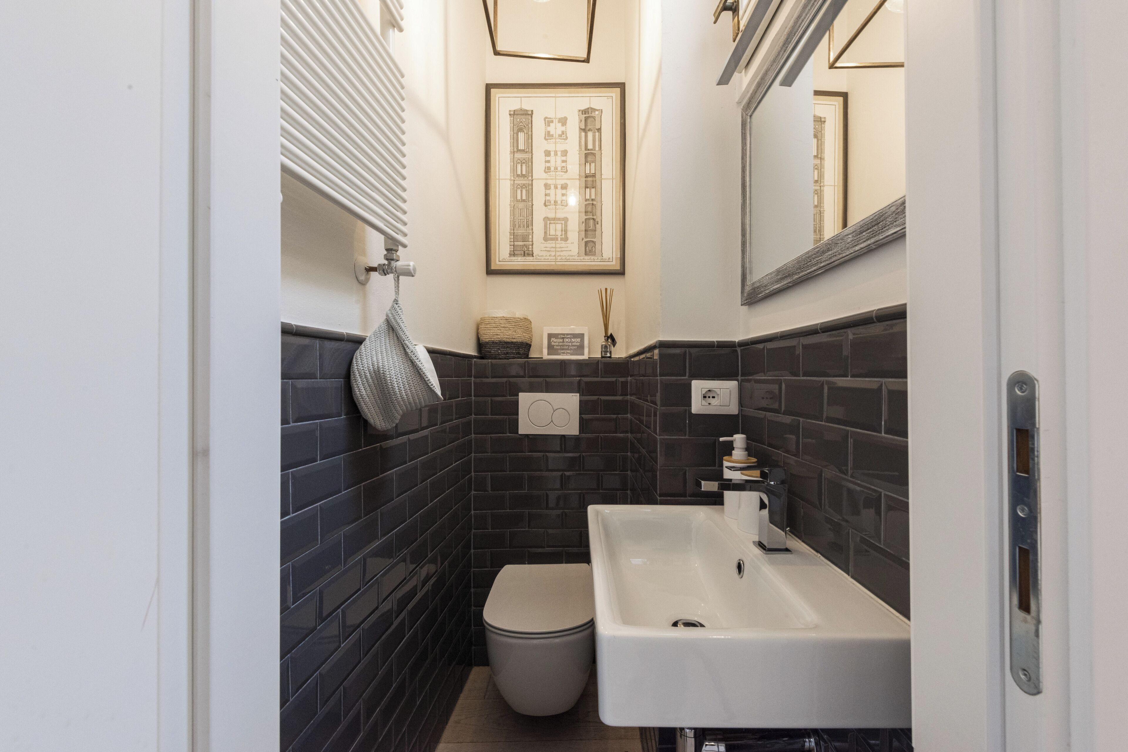 A bathroom with a white sink, dark tiled walls, a toilet, and a framed architectural drawing on the wall.