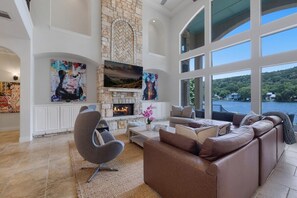 A spacious living room with a stone fireplace, comfortable seating, and large windows overlooking a body of water. - 4BR Lake Austin Estate | Private Dock + Wake Surf Water | 200 Ft Frontage (Austin)