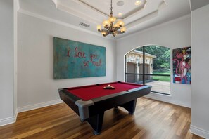 Uma mesa de bilhar em um salão com uma janela grande, um lustre e um quadro na parede.
