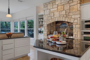 A kitchen with a stone fireplace, white cabinetry, and a breakfast bar with fruit and pastries. - 4BR Lake Austin Estate | Private Dock + Wake Surf Water | 200 Ft Frontage (Austin)