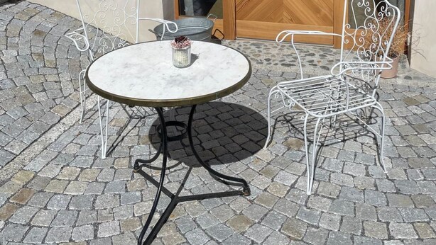 An outdoor seating area with a round table and two chairs, a wooden door with a chevron pattern, and a window with a view of the interior. - Stüvetta à Porta, (Scuol) by Interhome (Scuol)
