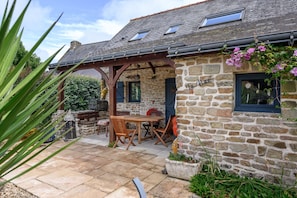 Ein Steinhaus mit einer Holzpergola, einer Steinsitzbank und einem Tisch mit Stühlen.