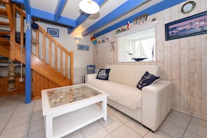 Una sala de estar con un sofá blanco, una mesa de centro de vidrio, una escalera de madera y un ventanal con vista a un velero.