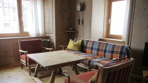 Een woonkamer met houten vloer, een geruit bankstel, een rode fauteuil, een houten salontafel en een open haard.
