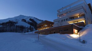 Ein modernes mehrgeschossiges Gebäude mit Holzfassade und Balkonen, gelegen in einer verschneiten Berglandschaft.
