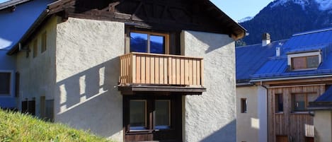 A two-story chalet with a wooden balcony, a stone facade, and a mountain backdrop.