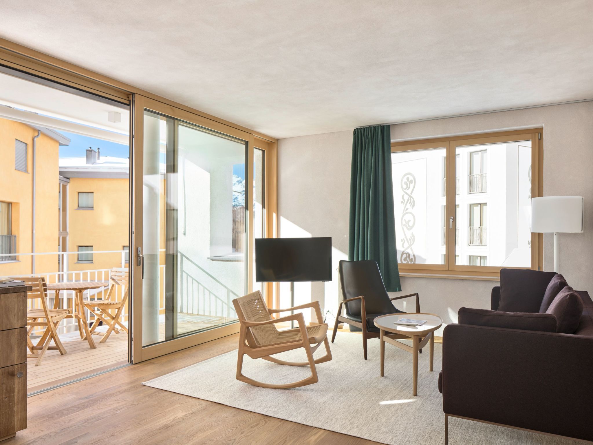 Ein modernes Wohnzimmer mit einer Couch, Sesseln, einem Fernseher und einem Balkon mit Holzmöbeln.