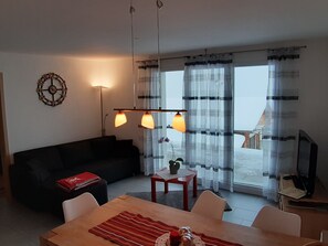 Ein Wohnzimmer mit einer Couch, einem Esstisch mit Stühlen, einem kleinen Tisch mit Pflanze, einer Wanduhr und einem großen Fenster mit Vorhängen.
