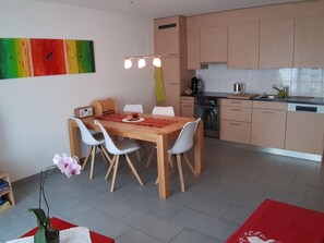 Una cocina compacta con electrodomésticos modernos, un comedor con una mesa de madera y sillas blancas, y un cuadro abstracto y colorido en la pared.