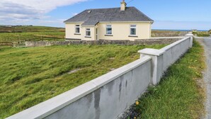 Una casa de un piso con techo gris y una pequeña chimenea, rodeada por un campo cubierto de hierba y un muro de hormigón.