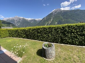 Un jardin avec un bac à fleurs en pierre et une haie, un ciel dégagé et une chaîne de montagnes en arrière-plan.