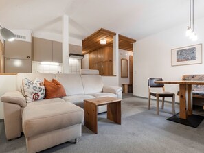 A modern living room with a sofa, a wooden coffee table, a dining table with chairs, and a kitchen area with cabinets and a refrigerator.