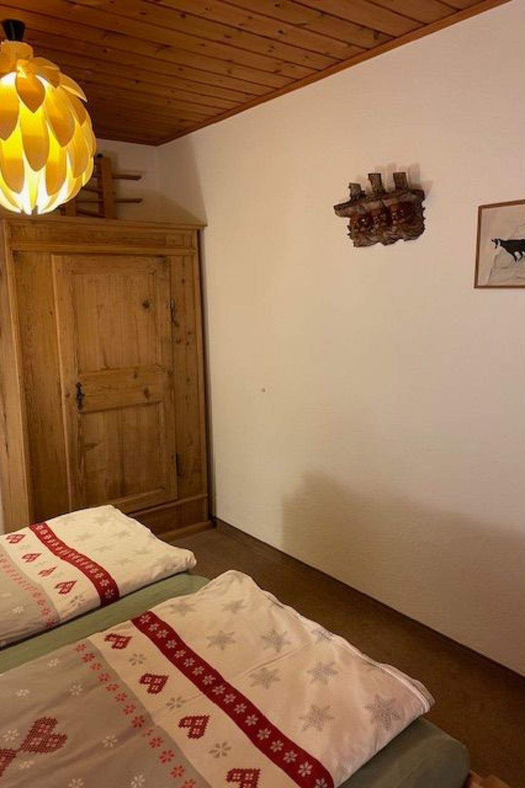 Quarto com guarda-roupa de madeira, cama com manta estampada, prateleira na parede com objetos decorativos e luminária pendente.