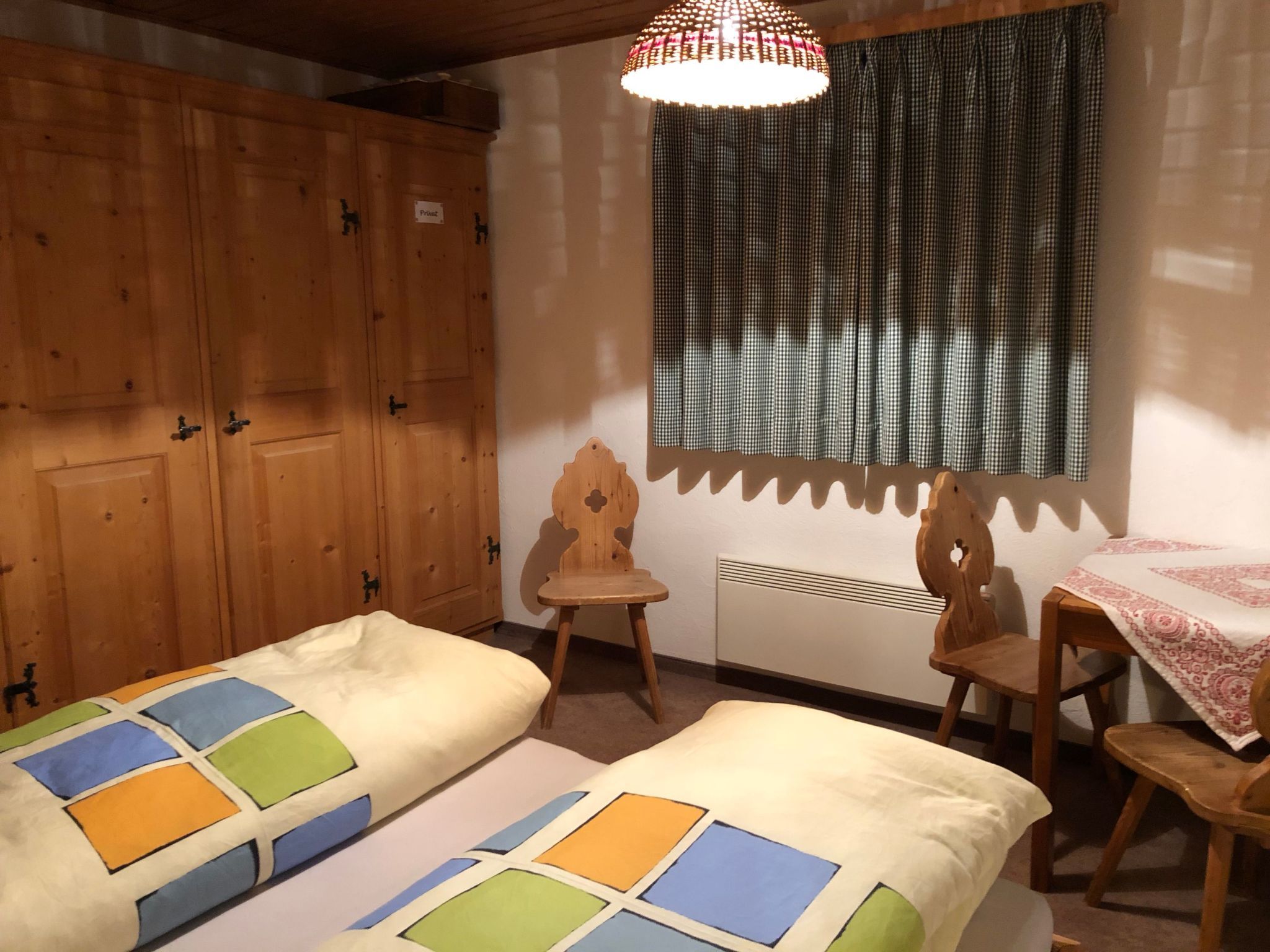 A room with two beds, a wooden wardrobe, a chair, a table with a tablecloth, and a window with curtains.
