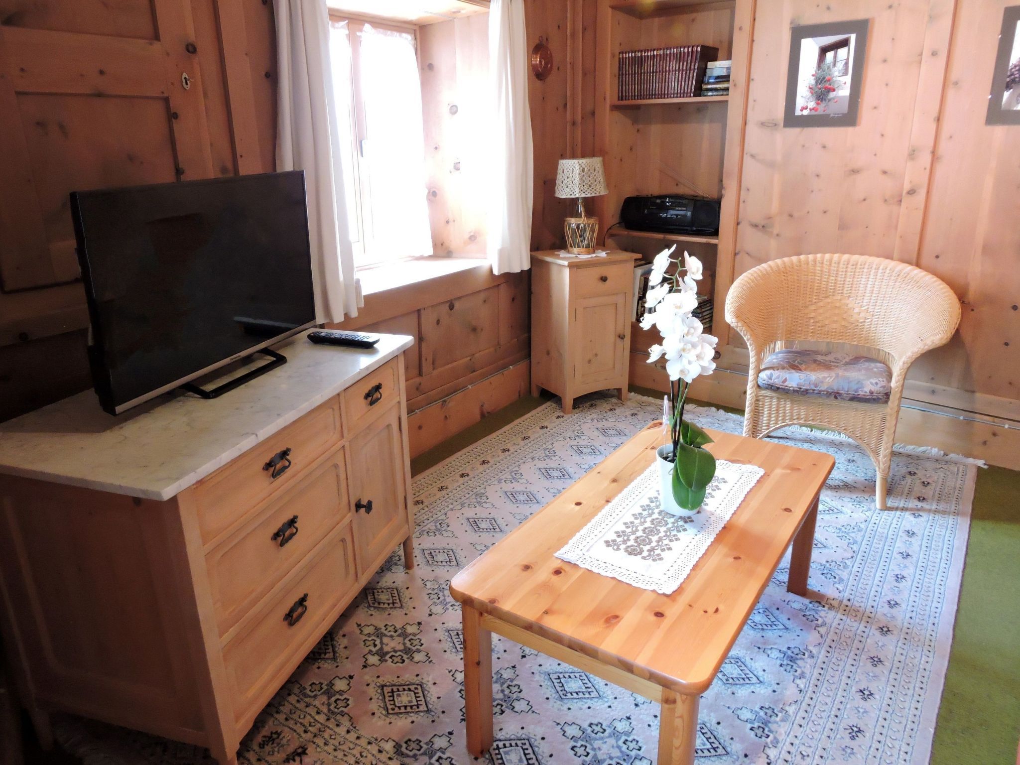 A wooden interior with a television on a desk, a wicker chair, a small table with a lace tablecloth, and patterned floor tiles.