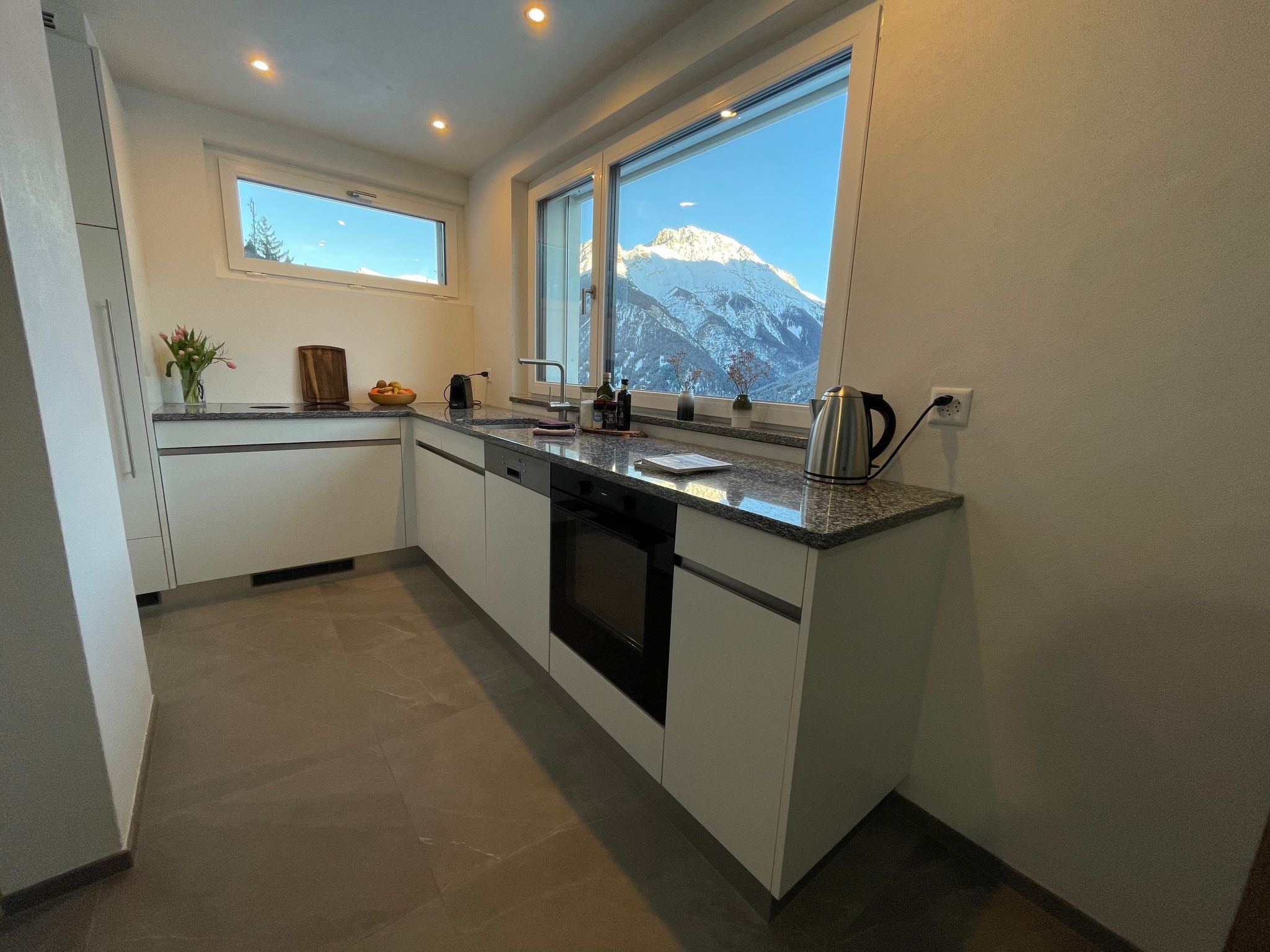 A modern kitchen with a large window, a kettle, and a bowl of fruit on the counter.