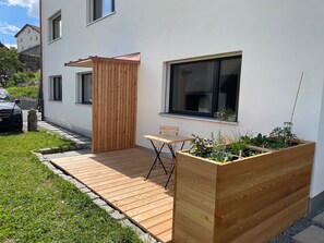 Eine Holzterrasse mit Tisch und Stühlen, ein erhöhter Blumenkasten mit Pflanzen sowie ein weißes Gebäude mit Fenstern.