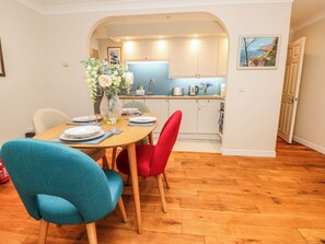 Comedor con mesa de madera dispuesta para cuatro, dos sillas, un florero con flores y una cocina al fondo.
