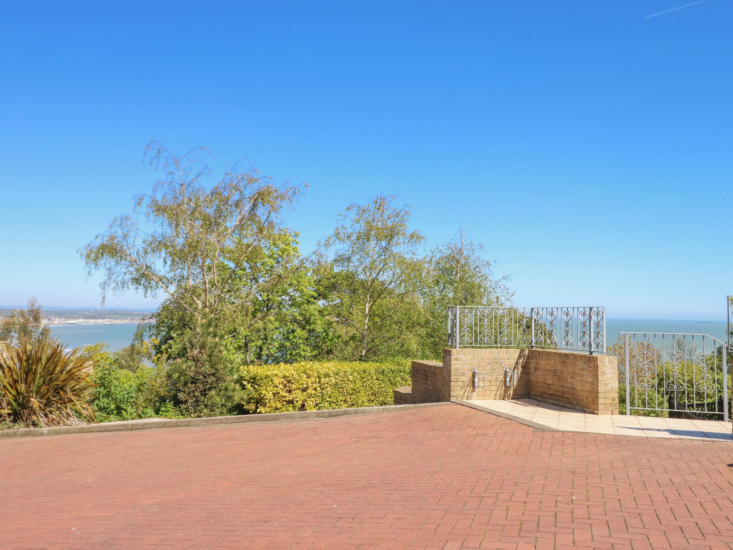 A paved outdoor area with a brick floor, surrounded by greenery and a view of the sea.