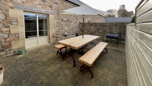 Een stenen gebouw met een patio, compleet met een houten tafel en banken, een barbecue en een grote parasol.
