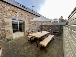 A stone building with a patio area featuring a wooden table and benches, a grill, and a large umbrella. - The Chimney (Bamburgh)