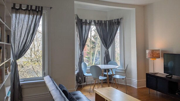 Ein Wohnzimmer mit einer Couch, einem Esstisch mit Stühlen, einem Fernseher und Vorhängen an den Fenstern.