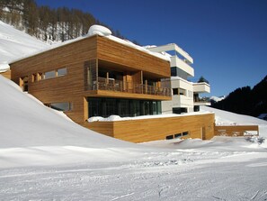 Ein modernes, mehrgeschossiges Gebäude mit Holzverkleidung, Balkonen und großen Fenstern, gelegen an einem verschneiten Hang.