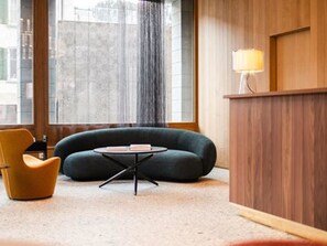 A room with a sofa, a chair, a small table, a wooden reception desk, and a large window. - Hotel Maistra 160, (Pontresina), by Interhome (Pontresina)