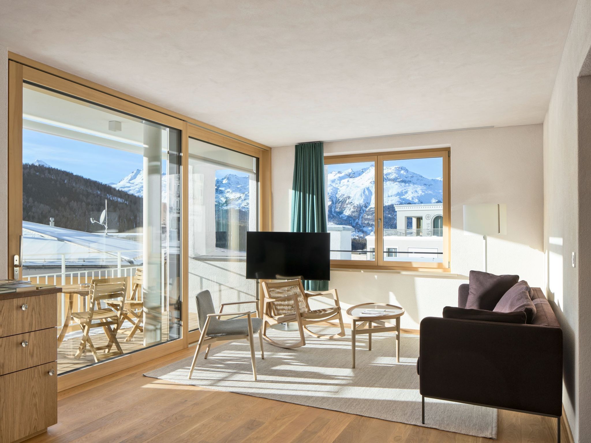 A modern living room with a large window, a flat-screen TV, wooden furniture, and a view of snow-capped mountains.