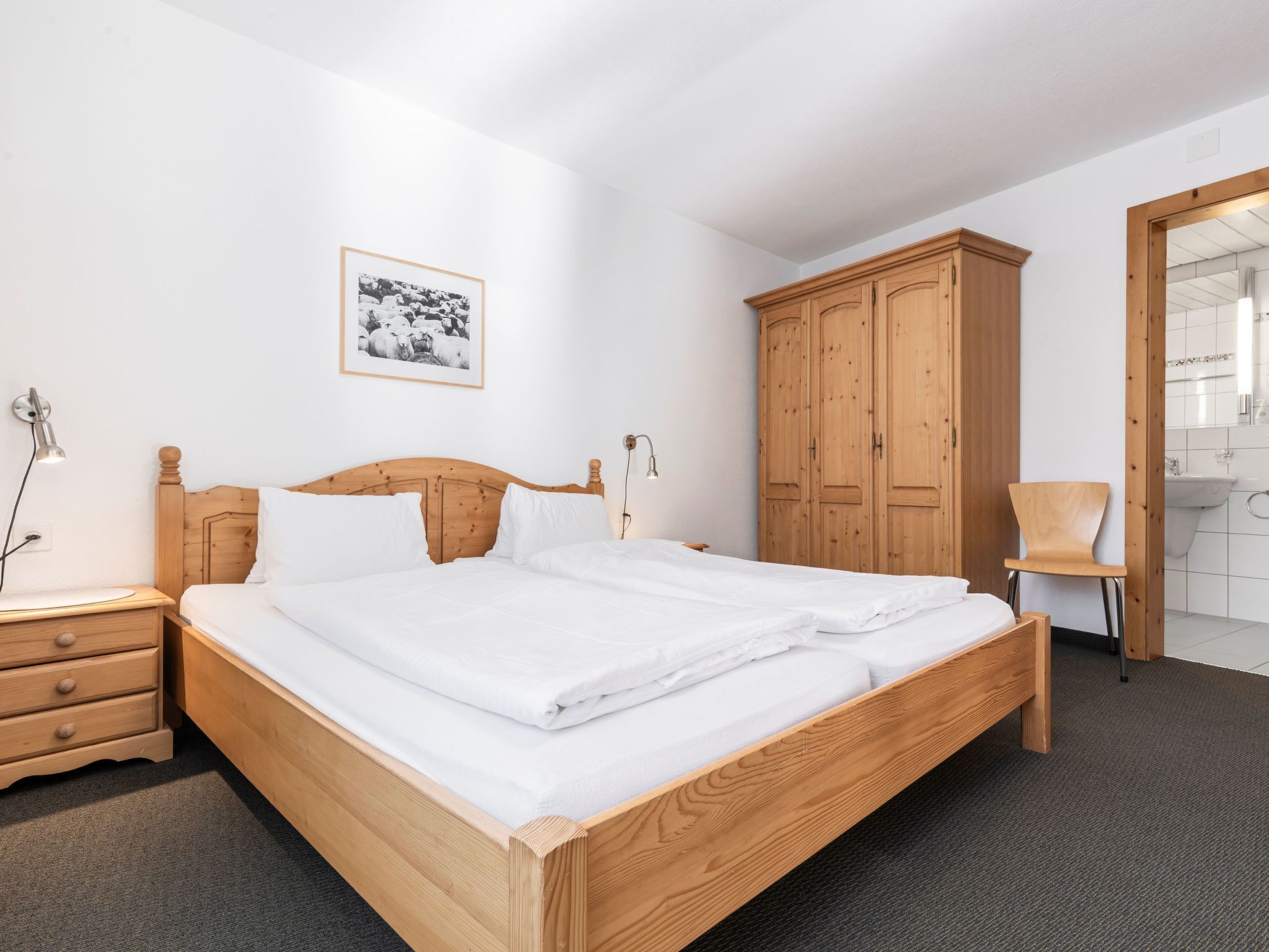 Un lit bien fait dans une chambre d’hôtel, avec une tête de lit en bois, une armoire en bois, une chaise et un tableau encadré accroché au mur.