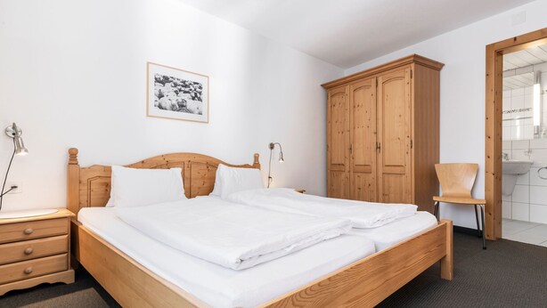 Ein ordentlich bezogenes Bett in einem Hotelzimmer, mit einem hölzernen Kopfteil, einem Schrank aus Holz, einem Sessel und einem gerahmten Bild an der Wand.