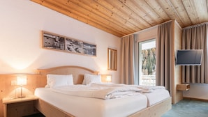 Quarto de hotel com cama de madeira, luminárias laterais, televisão de tela plana e uma janela grande com cortinas.