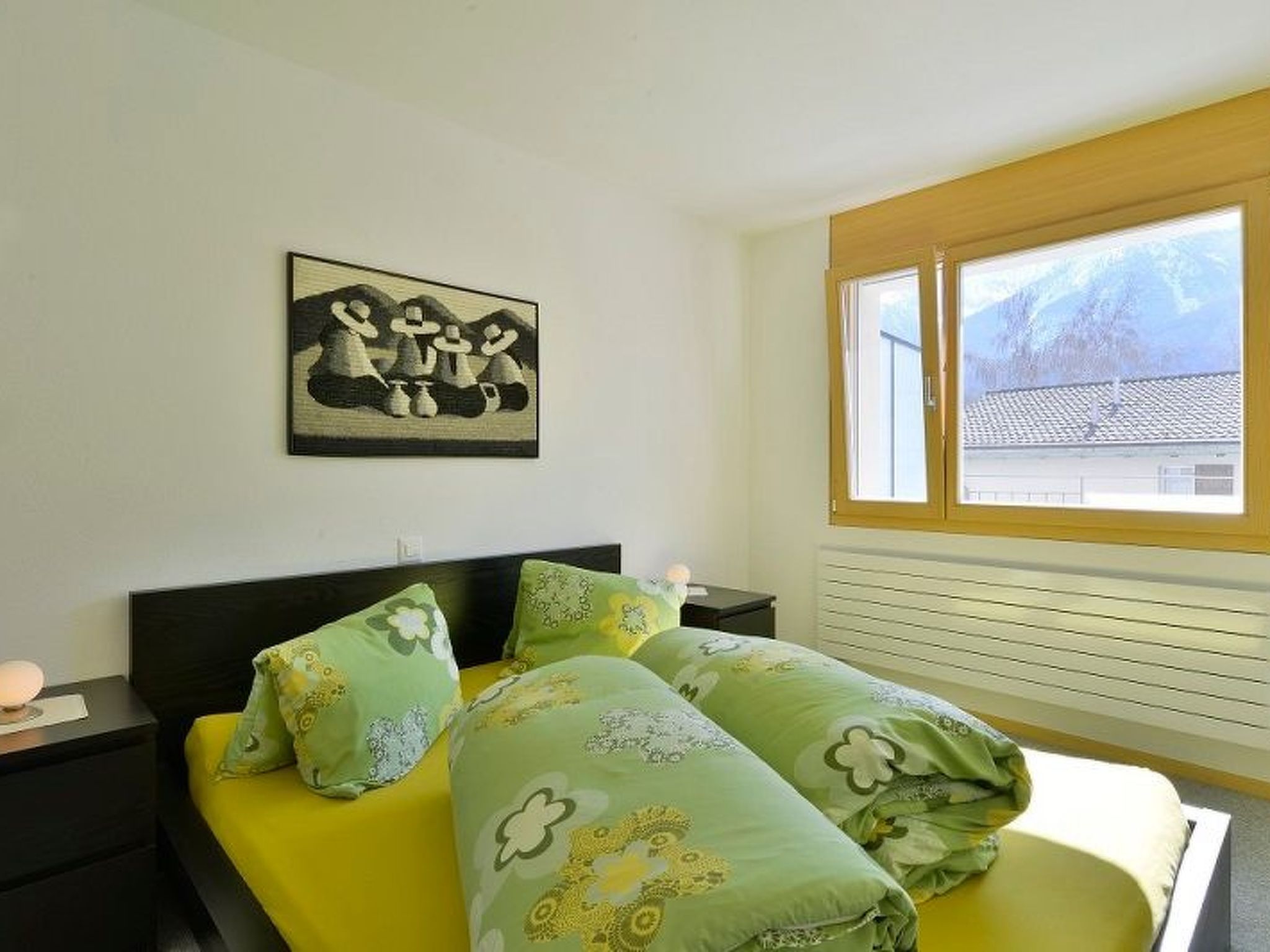 A bedroom with a bed, a nightstand, a lamp, a window with a view, and a painting on the wall.