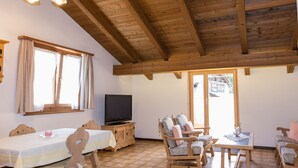 Ein Zimmer mit Holzbalkendecke, einem Fernseher, einem Esstisch mit Stühlen, einer Couch und einem Fenster mit Vorhängen.