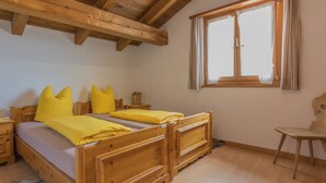 Ein Zimmer mit zwei Holzbetten, einem Vorhangfenster, einem Holzstuhl und einem kleinen Tisch mit Lampe.