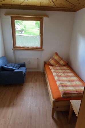 Une petite pièce comprenant un lit, une chaise et une fenêtre donnant sur un espace verdoyant.