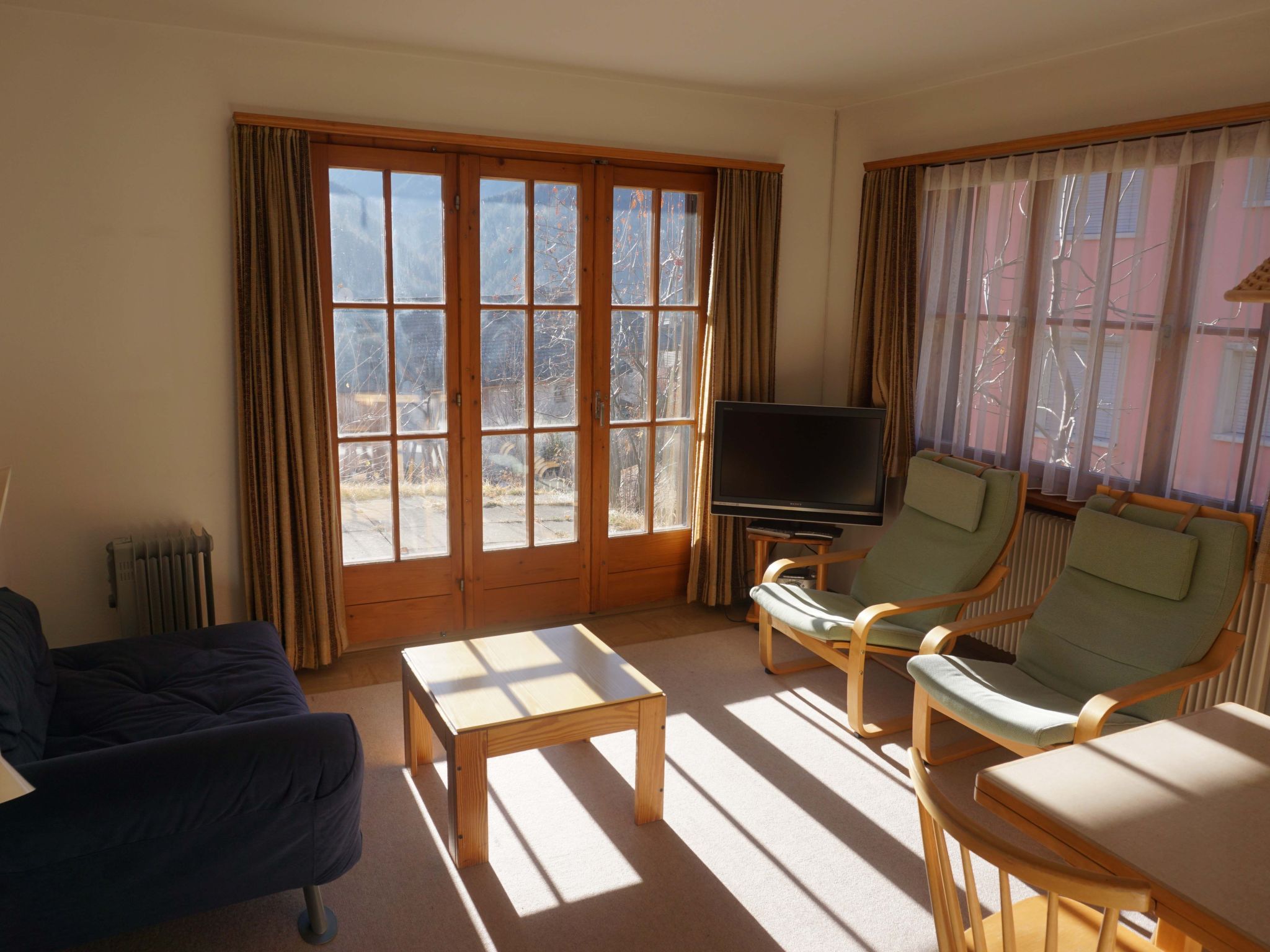 A living room with a sofa, two armchairs, a coffee table, a television, and large windows with curtains.