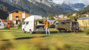 Un terrain de camping avec un camping-car blanc, une personne qui ajuste l’auvent et une chaîne de montagnes en arrière-plan.