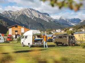 Un terrain de camping avec un camping-car blanc, une personne qui ajuste l’auvent et une chaîne de montagnes en arrière-plan.