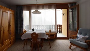 Ein Zimmer mit Esstisch, Stühlen, einer Couch, einem Kleiderschrank und einem großen Fenster mit durchsichtigen Vorhängen.