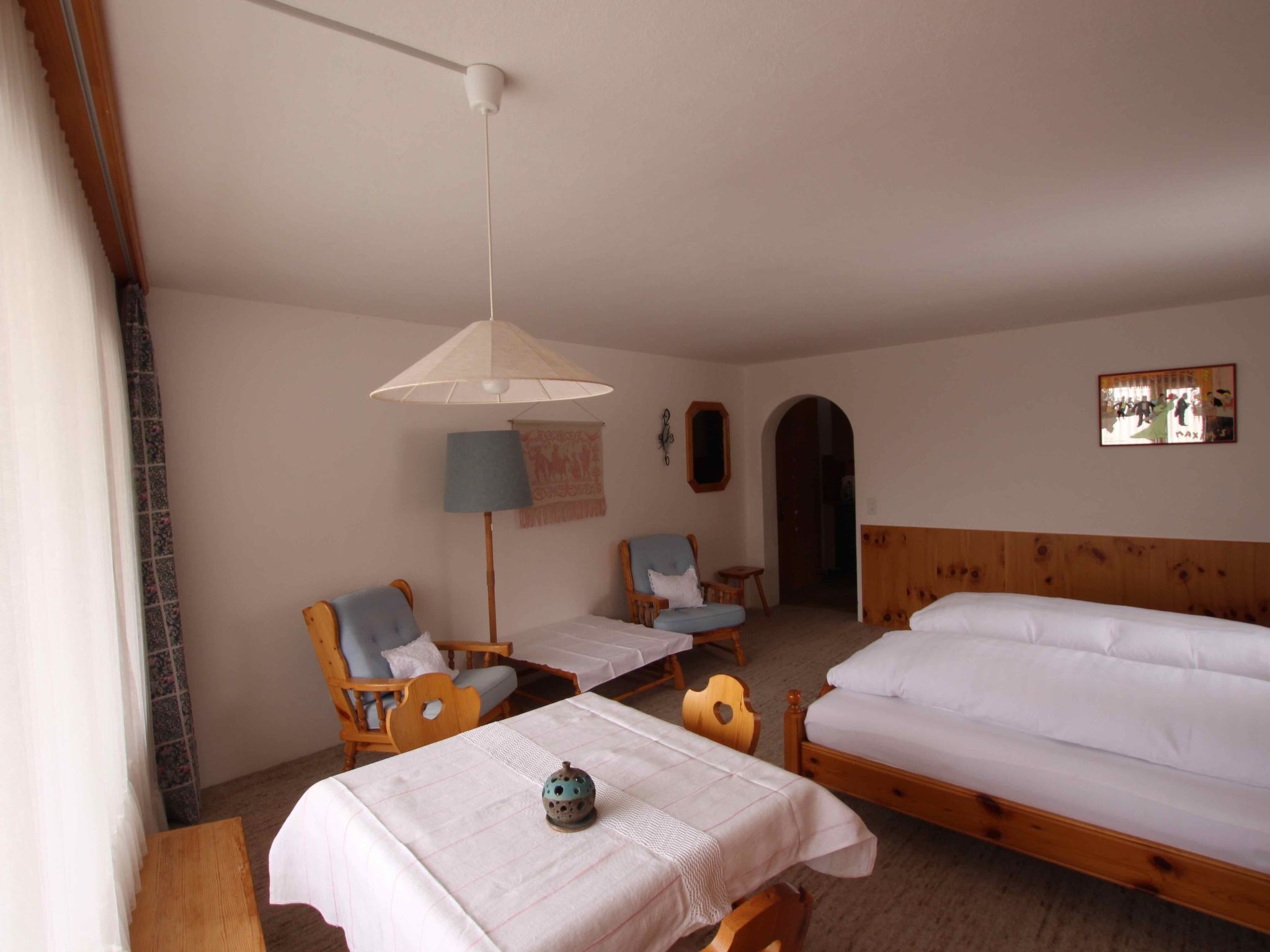 Ein Zimmer mit zwei Einzelbetten, einem Holz-Kopfstück, einem Tisch mit weißem Tuch, zwei Stühlen, einer Pendelleuchte und einem an der Wand hängenden Bild.