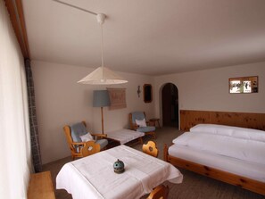 Ein Zimmer mit zwei Einzelbetten, einem Holz-Kopfstück, einem Tisch mit weißem Tuch, zwei Stühlen, einer Pendelleuchte und einem an der Wand hängenden Bild.