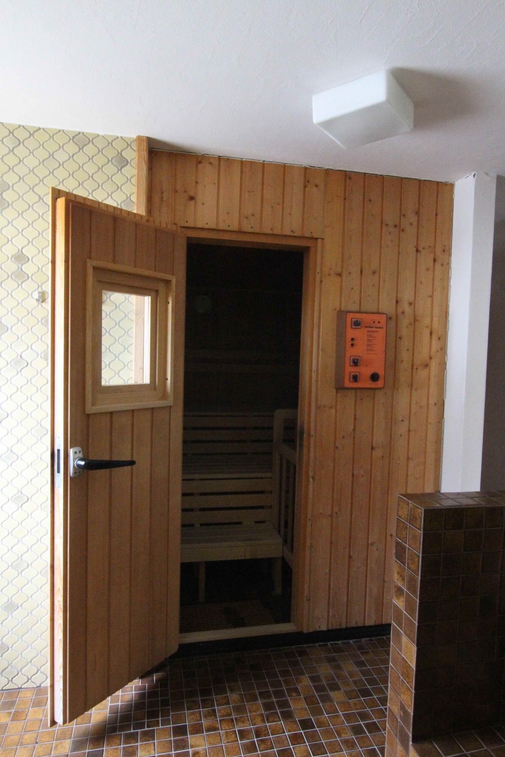 Une pièce avec une porte en bois, une petite fenêtre, un sol carrelé et un panneau de commande fixé au mur.