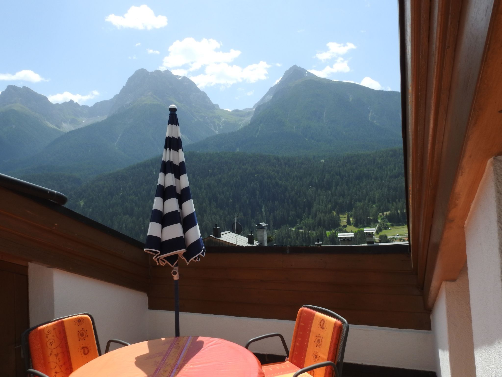 A balcony with a table, chairs, and a striped umbrella overlooking a mountainous landscape.