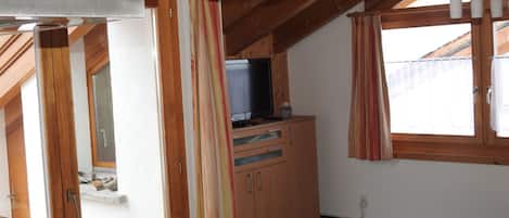 Une pièce avec un plafond en bois, un téléviseur posé sur un meuble et une fenêtre avec des rideaux.